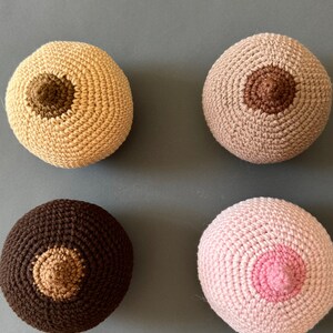 May include: Four crocheted breast shapes in different colors: beige, brown, pink, and dark brown. Each shape has a brown crocheted nipple in the center.