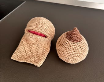 Op de afbeelding: Twee handgemaakte gehaakte items: een beige borst en een beige gezichtsmasker met een rode binnenkant. De borst heeft een bruine tepel. De items zijn gemaakt van garen en worden gepresenteerd op een donkerbruin oppervlak.