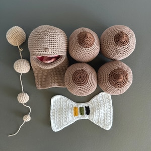 May include: A collection of handmade crochet items, including a beige face with a red interior, three pairs of crochet breasts, a white bow tie with green, yellow, and black stripes, and a string of beige crochet balls.