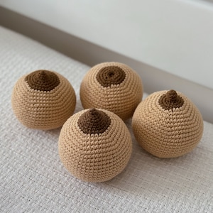 May include: Four crocheted breast-shaped objects. Each is a light tan color with a darker brown nipple. The objects are arranged on a white textured surface.