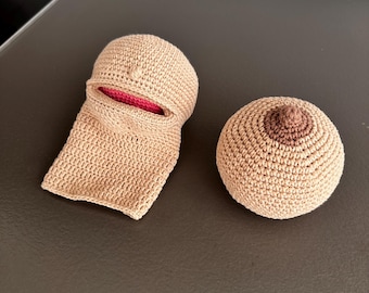 Op de afbeelding: Twee gehaakte items: een beige, openmondige hoofdbedekking met een rode binnenkant en een ronde, beige borst met een bruine tepel. De items zijn handgemaakt en liggen op een donkere ondergrond.
