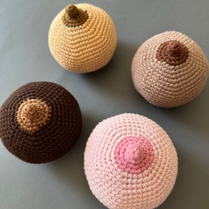 May include: Four crocheted breast shapes in different colors: beige, brown, pink, and light brown. Each shape has a crocheted nipple in the center.