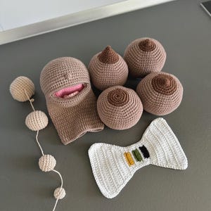May include: A collection of handmade crochet items, including a balaclava, breasts, a bow tie, and a string of small balls. The balaclava is tan with a pink mouth. The breasts are tan with brown nipples. The bow tie is white with colored stripes.