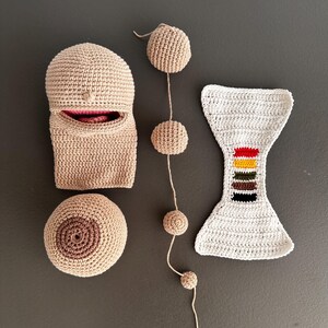 May include: A collection of handmade crochet items in beige and white. Includes a head-shaped piece with a pink interior, a round breast, a string of spheres, and a white hourglass-shaped piece with colorful stripes.