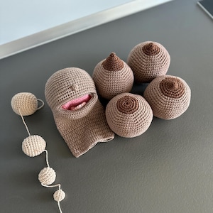 Op de afbeelding: Een verzameling handgemaakte gehaakte items, waaronder verschillende borstvormige vormen in verschillende maten en een gezichtsvormig stuk met een open mond. Er is ook een reeks kleine, ronde gehaakte ballen aanwezig. De items zijn in tinten beige en bruin.