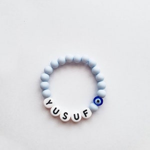 Könnte beinhalten: Ein hellblaues Perlenarmband mit dem Namen "YUSUF" in weißen Buchstabenperlen. Das Armband hat auch eine blaue böse-Augen-Perle, ein Schutzamulett.