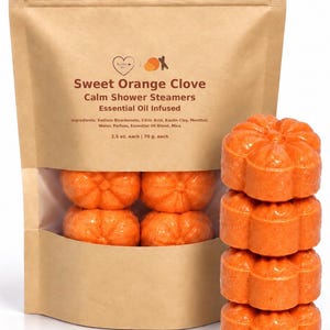 May include: A brown paper pouch labeled Sweet Orange Clove Calm Shower Steamers Essential Oil Infused, containing orange pumpkin-shaped shower steamers. A stack of four identical orange pumpkin-shaped shower steamers sits beside the pouch.