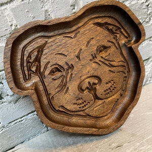 May include: A wooden tray shaped like a dog's head, with a carved design of a dog's face. The tray is made of dark brown wood and has a smooth, polished finish.