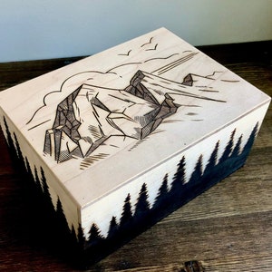 May include: A wooden box with a laser-engraved design of a mountain range and a forest on the sides. The top of the box is made of light wood and features a detailed drawing of a mountain peak.