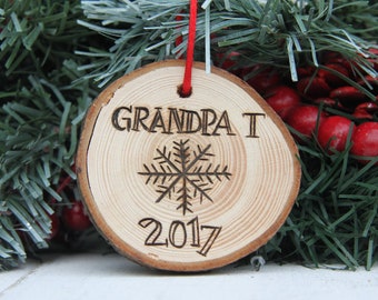 Może przedstawiać: Drewniana ozdoba choinkowa z napisem "GRANDPA T", wzorem płatka śniegu i rokiem "2017". Ozdoba wisi na czerwonym sznurku, na tle świątecznej zieleni i czerwonych jagód.