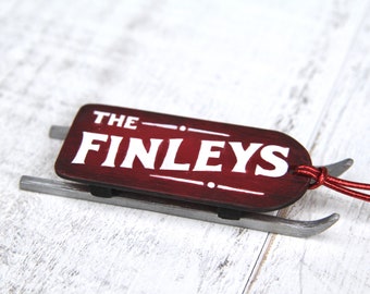 Puede incluir: Adorno de trineo de madera en miniatura con el texto "THE FINLEYS" en blanco sobre un fondo granate. El trineo tiene patines grises y un cordón rojo para colgar. Un artículo decorativo.