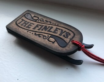 Puede incluir: Un adorno de madera con forma de trineo con el texto grabado "THE FINLEYS". El adorno está teñido de marrón con detalles en negro y un cordón rojo para colgarlo. El diseño presenta adornos decorativos.