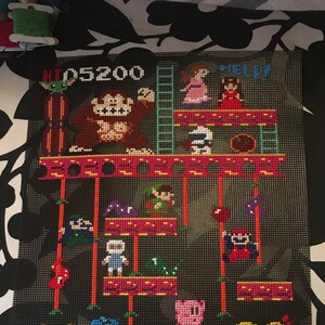 Puede incluir: Un dise&ntilde;o de punto de cruz pixelado con personajes de videojuegos populares, incluyendo Donkey Kong, Pac-Man y Pikachu. El dise&ntilde;o est&aacute; en un lienzo negro con un patr&oacute;n de cuadr&iacute;cula e incluye el texto "HI 05200" y "HELP".