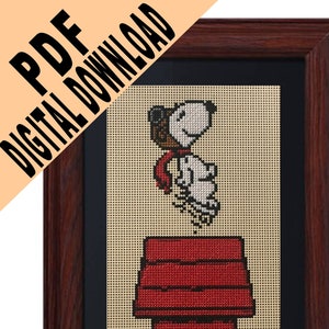 May include: Framed cross-stitch art featuring Snoopy, wearing a brown aviator cap and red scarf, jumping on top of his red doghouse. The artwork is set against a beige background and framed in dark wood. Text in the upper left corner reads "PDF DIGITAL DOWNLOAD."
