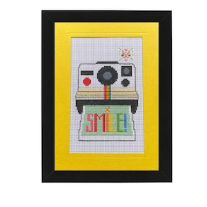May include: Framed cross-stitch art featuring a retro-style camera with a rainbow stripe and the word "SMILE!" in colorful letters. The artwork is set against a yellow background within a black frame, creating a vibrant and cheerful aesthetic.