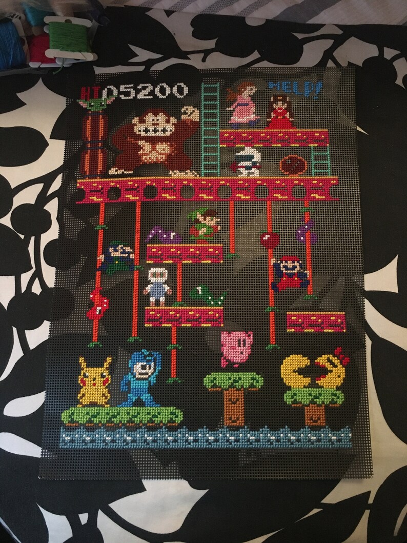 Puede incluir: Un dise&ntilde;o de punto de cruz pixelado con personajes de videojuegos populares, incluyendo Donkey Kong, Pac-Man y Pikachu. El dise&ntilde;o est&aacute; en un lienzo negro con un patr&oacute;n de cuadr&iacute;cula e incluye el texto "HI 05200" y "HELP".