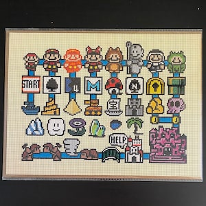 Op de afbeelding: Een kruissteekpatroon met verschillende kleurrijke gepixelde personages en objecten, waaronder een paddenstoel, een ster, een kasteel en een schedel. Het patroon is ingelijst in een beige lijst met een lichtbruine passe-partout.