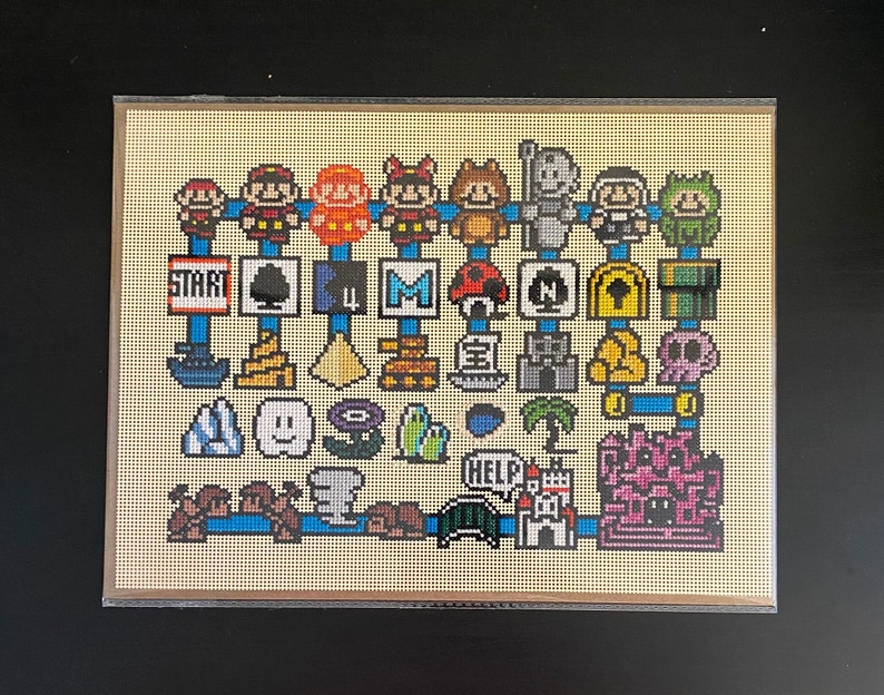 Op de afbeelding: Een kruissteekpatroon met verschillende kleurrijke gepixelde personages en objecten, waaronder een paddenstoel, een ster, een kasteel en een schedel. Het patroon is ingelijst in een beige lijst met een lichtbruine passe-partout.