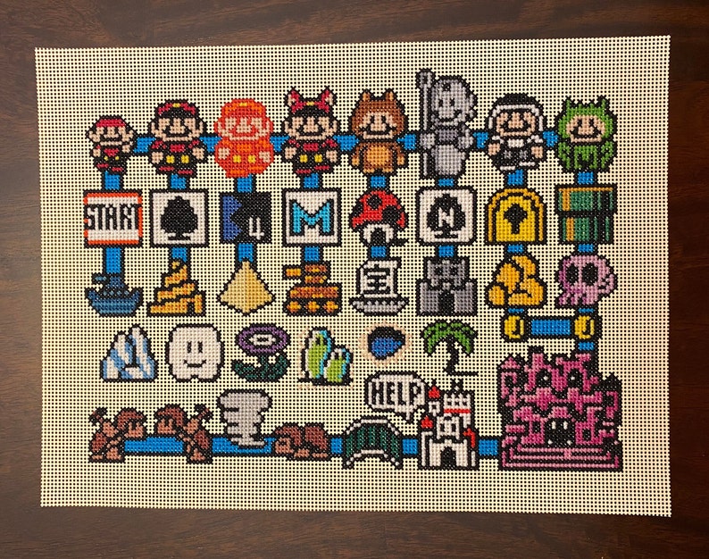 Op de afbeelding: Een kleurrijk kruissteekpatroon met personages en objecten uit de Super Mario Bros. videogame-serie. Het patroon bevat Mario, Luigi, Toad, Goomba's, een paddenstoel, een ster, een vraagtekenblok, een pijp en een kasteel.
