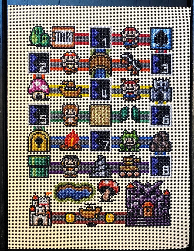May include: A framed needlepoint depicting a pixelated video game board with various characters and elements. The board includes numbers, a start marker, and iconic game imagery in a grid-like pattern. The colour palette includes red, blue, green, and yellow.