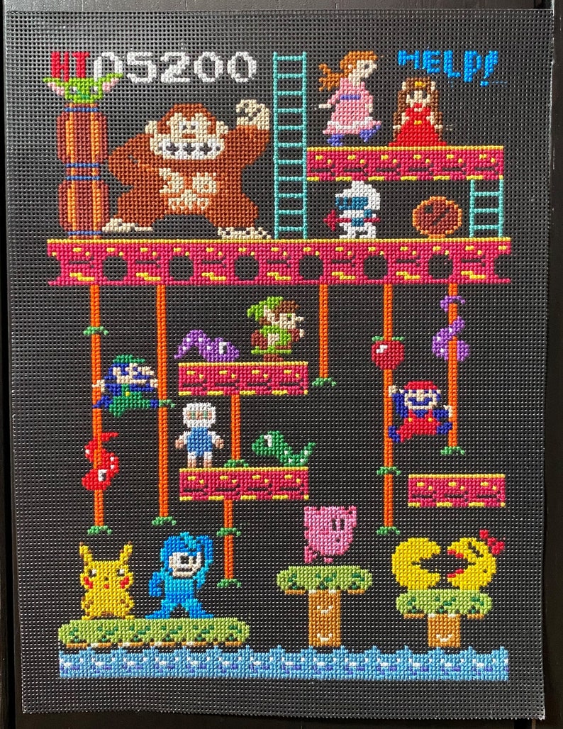 Puede incluir: Un dise&ntilde;o de punto de cruz pixelado con personajes de videojuegos populares. El dise&ntilde;o incluye un gorila marr&oacute;n grande, un robot azul, un Pikachu amarillo, un Kirby rosa y un personaje verde de Mario. El texto "HI 05200 HELP?" se muestra en la parte superior del dise&ntilde;o.