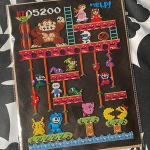 Puede incluir: Un patr&oacute;n de punto de cruz que representa una escena cl&aacute;sica de un videojuego. La escena presenta un gran gorila marr&oacute;n, una escalera y varios personajes, entre ellos un cerdo rosa, un p&aacute;jaro amarillo y un robot azul. El texto "HI 05200 HELP!" se muestra en la parte superior del patr&oacute;n.