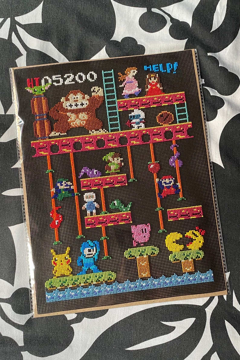 Puede incluir: Un patr&oacute;n de punto de cruz que representa una escena cl&aacute;sica de un videojuego. La escena presenta un gran gorila marr&oacute;n, una escalera y varios personajes, entre ellos un cerdo rosa, un p&aacute;jaro amarillo y un robot azul. El texto "HI 05200 HELP!" se muestra en la parte superior del patr&oacute;n.