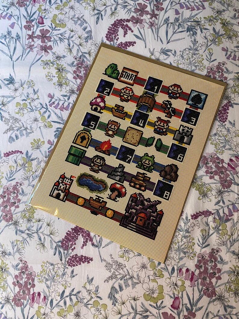 May include: A framed cross-stitch depicting a pixelated design inspired by a classic video game. The artwork includes iconic characters, level elements, and the word "START". The colour palette includes red, green, blue, brown, and yellow.