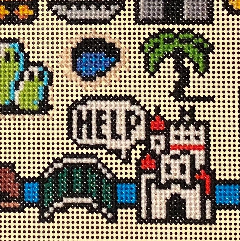 Op de afbeelding: Een kruissteekpatroon met een wit kasteel, een groene brug, een blauw meer, een palmboom en een spreekballon met de tekst "HELP".
