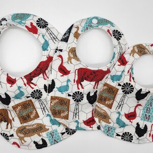 May include: Three white cotton bibs with a farm animal print. The bibs have a white background with a repeating pattern of red and black cows, brown pigs, black and white chickens, blue ducks, and brown windmills. The bibs have a white border and a white neck opening. The bibs have the text "Farm Sweet Farm" and "Farm Fresh Everyday" printed on them.