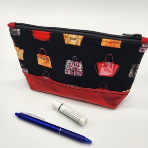 May include: A red and black zippered pouch with a black fabric lining and a red faux leather bottom. The pouch is decorated with a repeating pattern of colorful handbags.