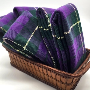 May include: A woven basket filled with folded purple, green, and white plaid fabric. The fabric has a herringbone weave and is neatly arranged in the basket. The basket is made of brown wicker.