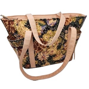 May include: A large floral tote bag with brown straps. The bag is made of a tapestry fabric with a floral pattern in shades of green, yellow, brown, and purple.
