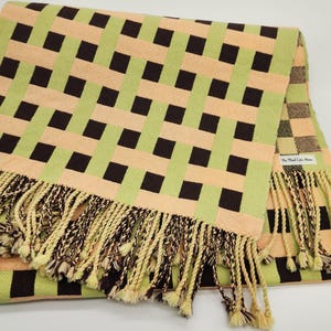 May include: A woven blanket with a geometric pattern of brown squares on a peach and light green background. The blanket has a fringe of cream and brown yarn along the edges. The tag reads "The Plaid Cake Home".