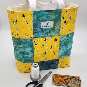May include: A yellow and teal tote bag with a patchwork design featuring kites. The bag has white handles and a label that reads "The Plaid Cat's Meow".