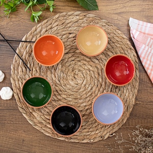 May include: Set of six small ceramic bowls in various colours, including orange, red, green, black, beige, and lavender. The bowls are arranged in a circle on a woven straw mat.