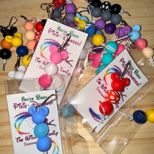 May include: Assorted colorful Mickey Mouse-themed keychains. Each keychain features a Mickey Mouse head design and multiple round beads in various colors. Some keychains are packaged with cards that read "You've Been Pixie-Dusted!"