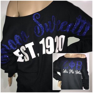 May include: Black off-the-shoulder sweatshirt with blue glitter text that reads "Sweet" and "EST. 1920". A smaller image shows the text "ZΦB Beta Phi Beta" in blue glitter and white.