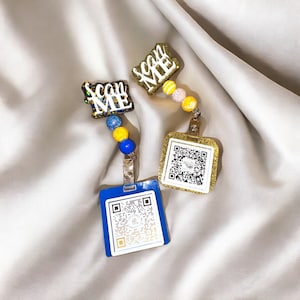 May include: Two blue and gold keychains with the words "Scan Me" on them. Each keychain has a blue and yellow beaded chain and a blue card with a QR code.