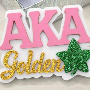 Puede incluir: Un emblema decorativo con las letras "AKA" en rosa, y la palabra "Golden" en escritura dorada con purpurina. También hay una hoja verde con purpurina. El diseño está sobre un fondo blanco.