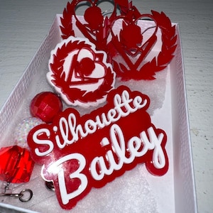 Könnte beinhalten: Eine Sammlung roter und weißer Acryl-Accessoires, darunter Ausweisrollen und dekorative Formen. Die Ausweisrollen tragen den Namen "Silhouette Bailey" in weißer Schrift auf rotem Hintergrund. Weitere Formen sind Herzen und Lorbeerkränze.