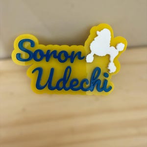 May include: A yellow and blue Soron Udechi poodle-shaped pin. The pin features the name "Soron Udechi" in blue cursive lettering, with a white poodle silhouette on the right side. The pin is set against a light wooden background.