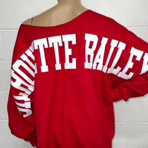 May include: Red sweatshirt with white text that reads "SQUEETIE BAILEY" on the back. The text is in a large, bold font and is arched across the back of the sweatshirt.