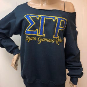 May include: Navy blue off-the-shoulder sweatshirt with the Greek letters ΣΓΡ in blue glitter and gold glitter lettering. The words "Sigma Gamma Rho" are written below the letters in gold glitter.