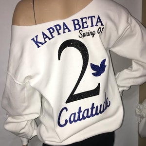 Peut inclure: Sweat-shirt blanc à épaules dénudées avec un texte en paillettes bleues qui dit "Kappa Beta Spring 01" et un grand chiffre "2" en paillettes noires et bleues avec un oiseau en paillettes bleues au-dessus. En dessous du chiffre, le texte "Catatude" en paillettes bleues.