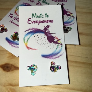May include: White cards with the text "Magic is Everywhere" and a silhouette of a fairy with a rainbow trail. Two Mickey Mouse-shaped sequin embellishments are visible. The cards are on a wooden surface.