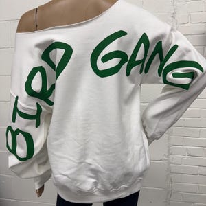 Op de afbeelding: Witte off-shoulder sweatshirt met groene letters die "Big Gang" op de mouwen lezen.