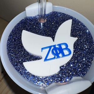 Può includere: Un bicchiere bianco con una base di glitter blu e una colomba bianca con le lettere "ZΦB" in blu. La colomba è attaccata al bicchiere con una cannuccia di plastica trasparente.