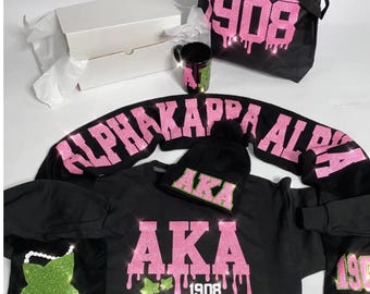 Op de afbeelding: Een verzameling zwarte kleding en accessoires met roze en groene glitteraccenten. Items zijn onder meer een totebag, sweatshirt, beanie, mok en een shirt met lange mouwen, allemaal met de tekst "Alpha Kappa Alpha" en "1908". Er is ook een witte doos aanwezig.