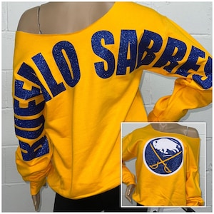 May include: Yellow off-the-shoulder sweatshirt with blue glitter lettering that says "Buffalo Sabres" on the back and a blue and white Buffalo Sabres logo on the front.